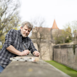 Eine Person in kariertem Hemd führt an der Nürnberger Stadtmauer eine Inspektion oder Probenahme durch; im Hintergrund sind Bäume sowie ein Teil der historischen Mauer mit Turm zu sehen.