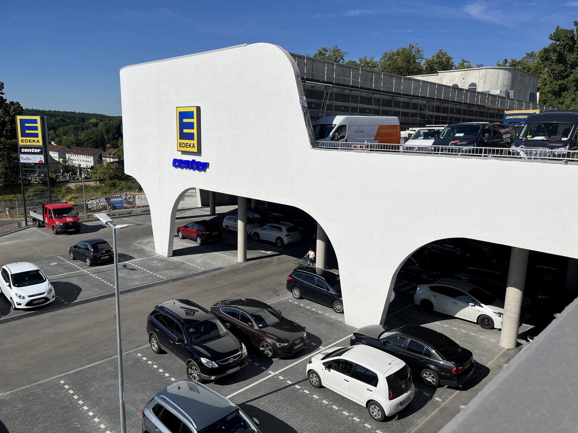 Parkbereich eines EDEKA-Centers mit zweistöckiger Parkstruktur, weißen Bögen und mehreren geparkten Autos; EDEKA-Logo und Beschilderung sichtbar, im Hintergrund Bäume und weitere Gebäude bei klarem Himmel.
