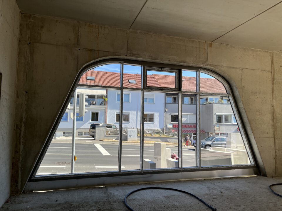 Großes Fenster mit geschwungener Oberkante in einem Betonbau; Blick auf eine Straße mit geparkten Autos, Passanten und gegenüberliegenden Wohnhäusern mit blauen Fassaden und roten Ziegeldächern; Innenraum im Rohbauzustand.