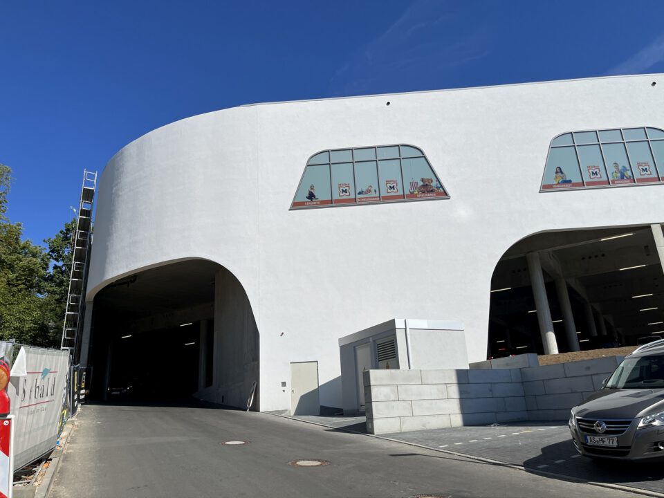 Modernes Gebäude mit geschwungener weißer Fassade und zwei großen Torbögen im Erdgeschoss; darüber Fenster mit Werbebeklebung, daneben eine Straße mit geparktem Auto, Baugerüst und Absperrmaterialien.