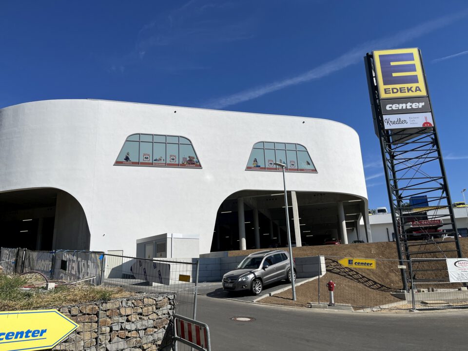 Modernes, weißes Gebäude mit großen, auffällig geformten Fenstern; rechts im Bild ein Schild mit der Aufschrift ‚EDEKA center‘. Vor dem Gebäude befinden sich Bauzäune und Materialien, was auf laufende oder kürzlich abgeschlossene Bauarbeiten hinweist.