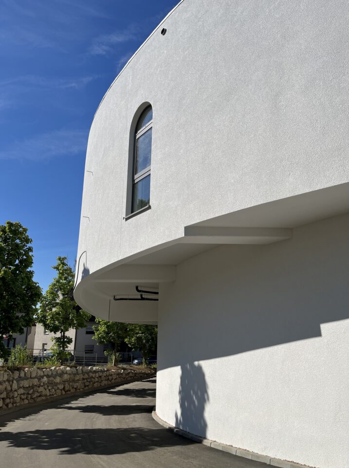 Weißes, geschwungenes Gebäude mit glatter Fassade und einem einzelnen, bogenförmigen Fenster im oberen Bereich; daneben ein Weg mit Bäumen und einer niedrigen Steinmauer, unter klarem, blauem Himmel.