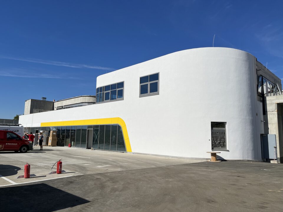 Modernes, weißes Gebäude mit geschwungener Architektur, großen Fenstern im Obergeschoss und einer Reihe verglaster Eingangstüren mit gelbem Akzent im Erdgeschoss. Davor befinden sich mehrere Personen, ein roter Transporter sowie ein offener Parkplatz mit zwei Feuerlöschern auf dem Boden.