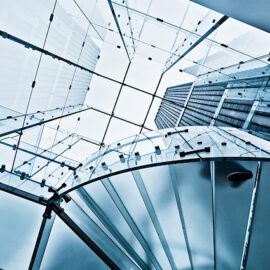 Das Bild zeigt eine moderne Wendeltreppe aus Glas und Metall, von unten nach oben fotografiert. Durch die Treppe hindurch sind ein Glasdach und hohe Gebäude im Hintergrund sichtbar.