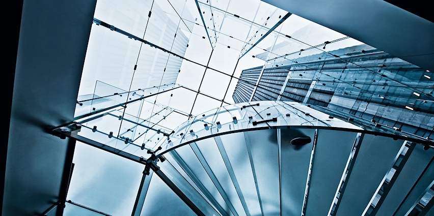 Das Bild zeigt eine moderne Wendeltreppe aus Glas und Metall, von unten nach oben fotografiert. Durch die Treppe hindurch sind ein Glasdach und hohe Gebäude im Hintergrund sichtbar.