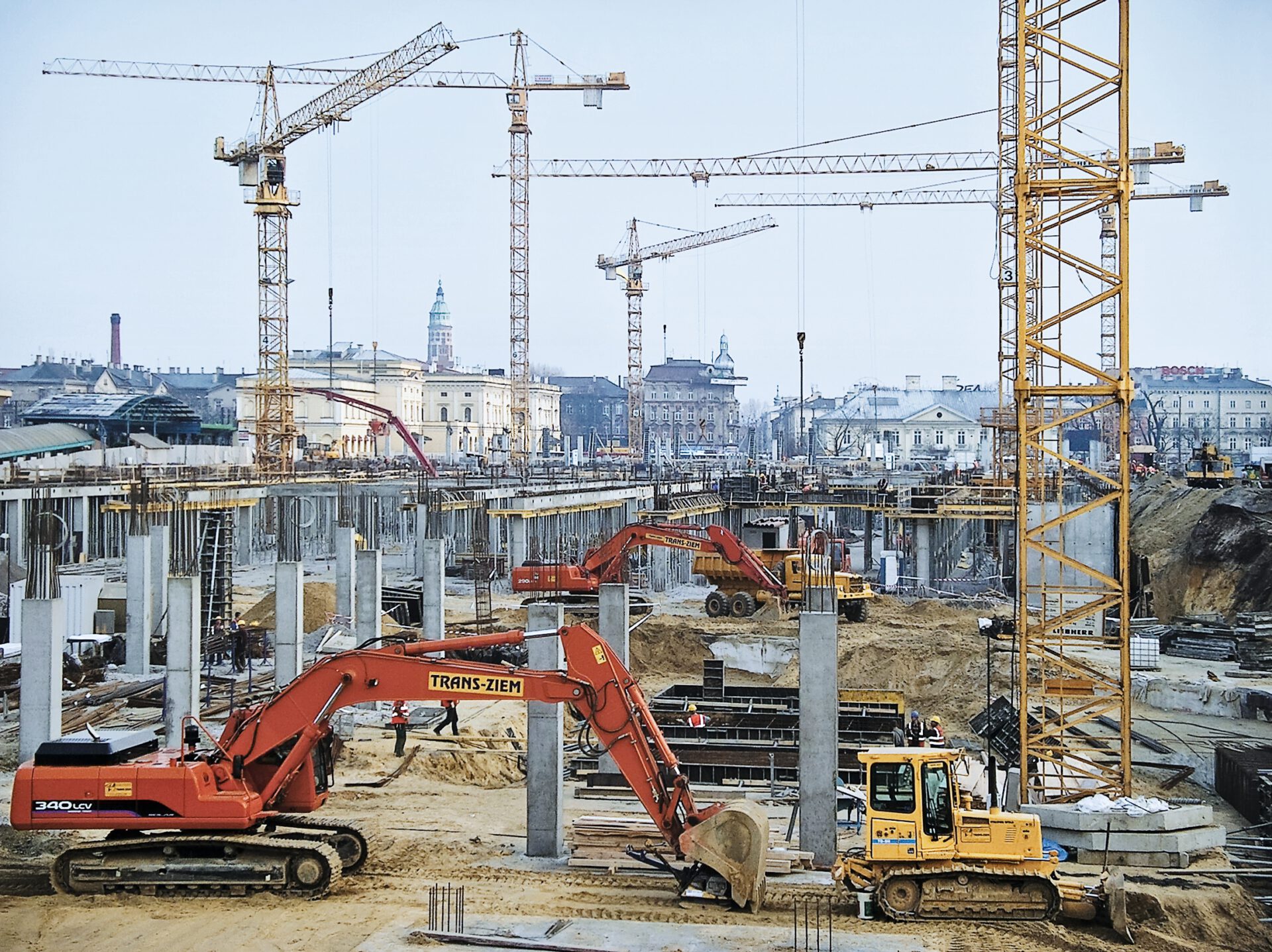 Das Bild zeigt eine große Baustelle mit mehreren Kränen und schweren Baumaschinen. Im Vordergrund arbeiten zwei Bagger, einer rot, der andere gelb, umgeben von Betonpfeilern und Trägern inmitten eines städtischen Umfelds.