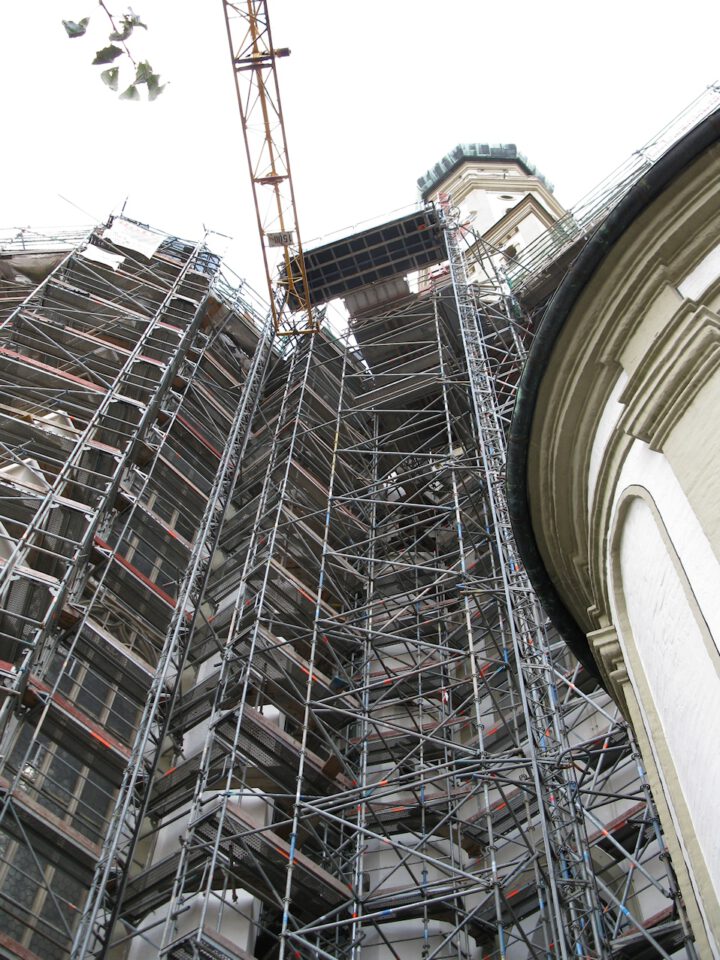 Gebäude mit mehrstöckigem Metallgerüst vollständig eingerüstet, ein Kran ragt über das Gerüst hinaus, im Hintergrund ist ein Teil eines Turms oder einer Spitze zu sehen.