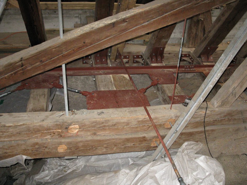 Holzkonstruktion mit mehreren Balken, die durch Metallplatten und -winkel verbunden sind, zusätzlich stabilisiert durch Metallstangen und Seile; im Hintergrund sind Werkzeuge und Baumaterialien zu sehen.