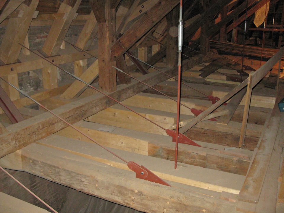 Innenansicht einer hölzernen Dachkonstruktion mit Balken und Fachwerk, zusätzlich stabilisiert durch Metallstangen und Spannschlösser.