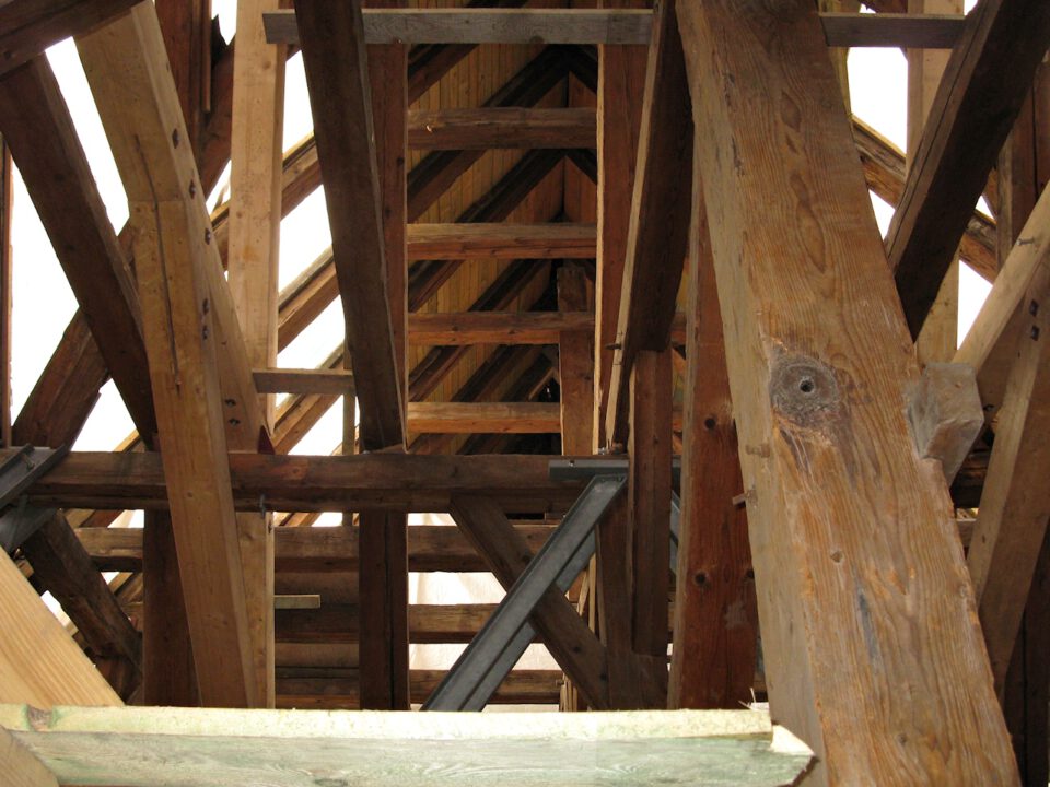 Innenansicht einer hölzernen Dachkonstruktion mit sichtbaren Balken und Fachwerk in Dreiecksform, verstärkt durch Metallwinkel; das Holz ist naturbelassen mit sichtbarer Maserung.