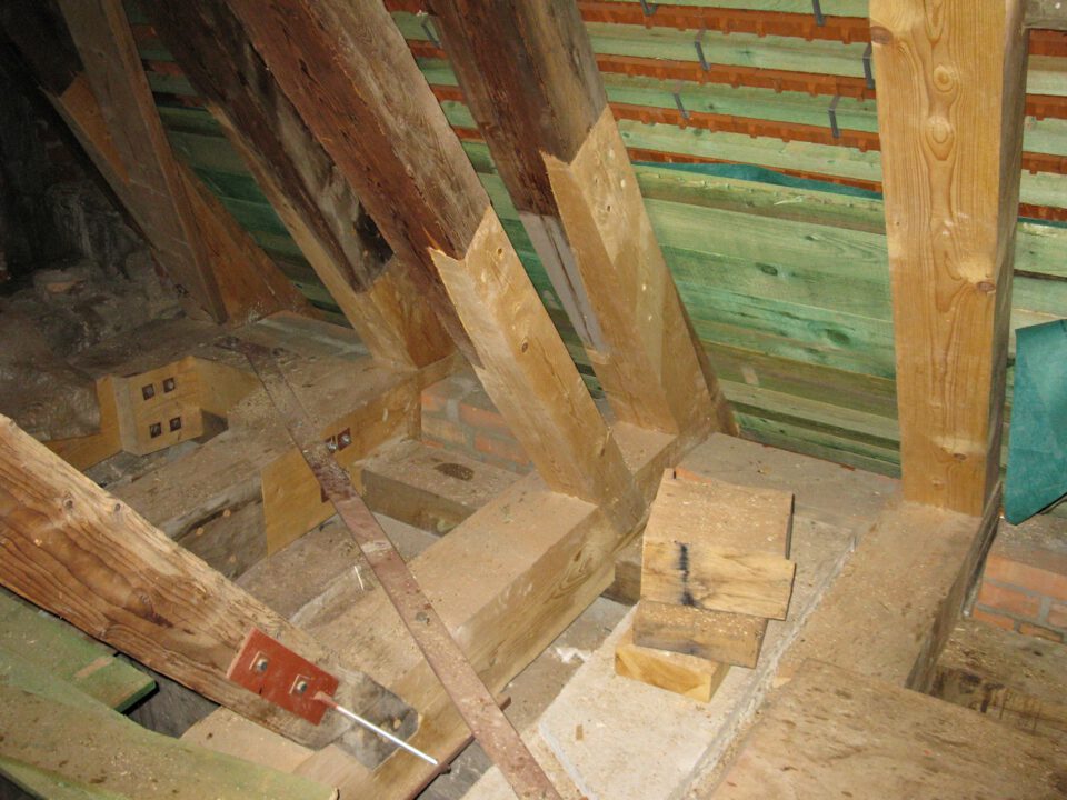 Innenansicht einer Dachkonstruktion mit mehreren Holzbalken und Verstärkungen, verbunden durch Metallwinkel und Schrauben; die Wände bestehen aus Ziegelmauerwerk, eine Seite ist mit grünem Holz verkleidet.