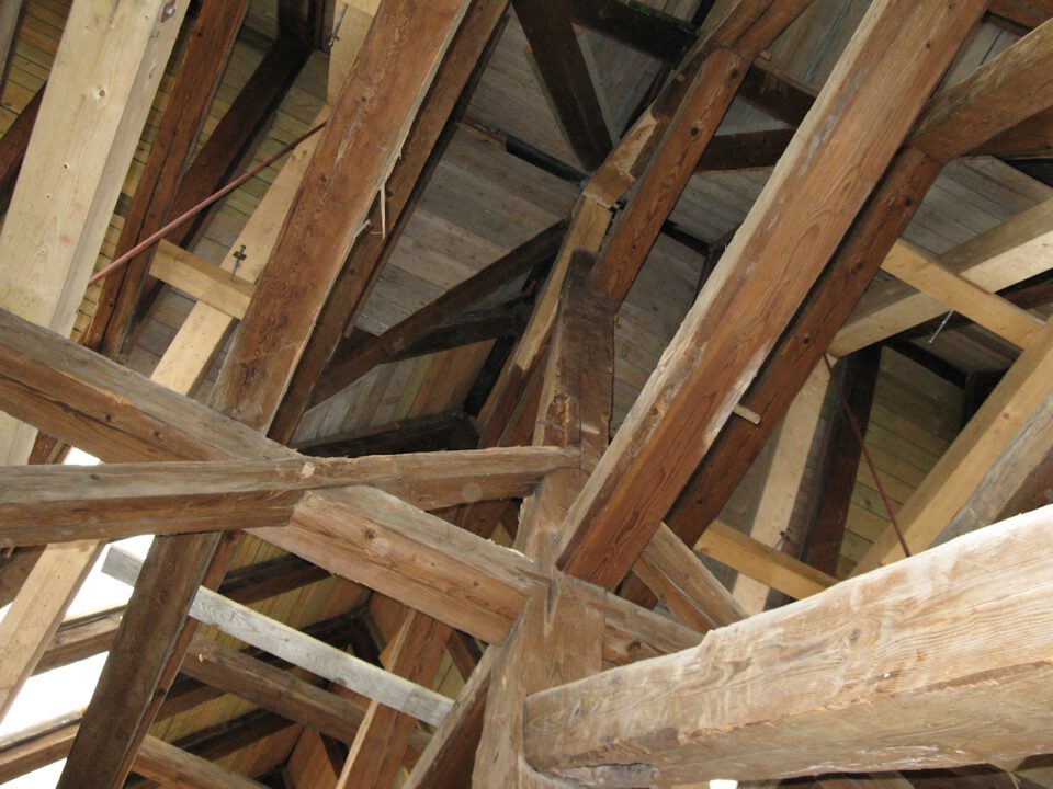 Nahaufnahme von hölzernen Dachbalken und Fachwerk, die sich in verschiedenen Winkeln kreuzen und die Dachkonstruktion tragen; das Holz wirkt gealtert mit sichtbarer Maserung und Rissen.