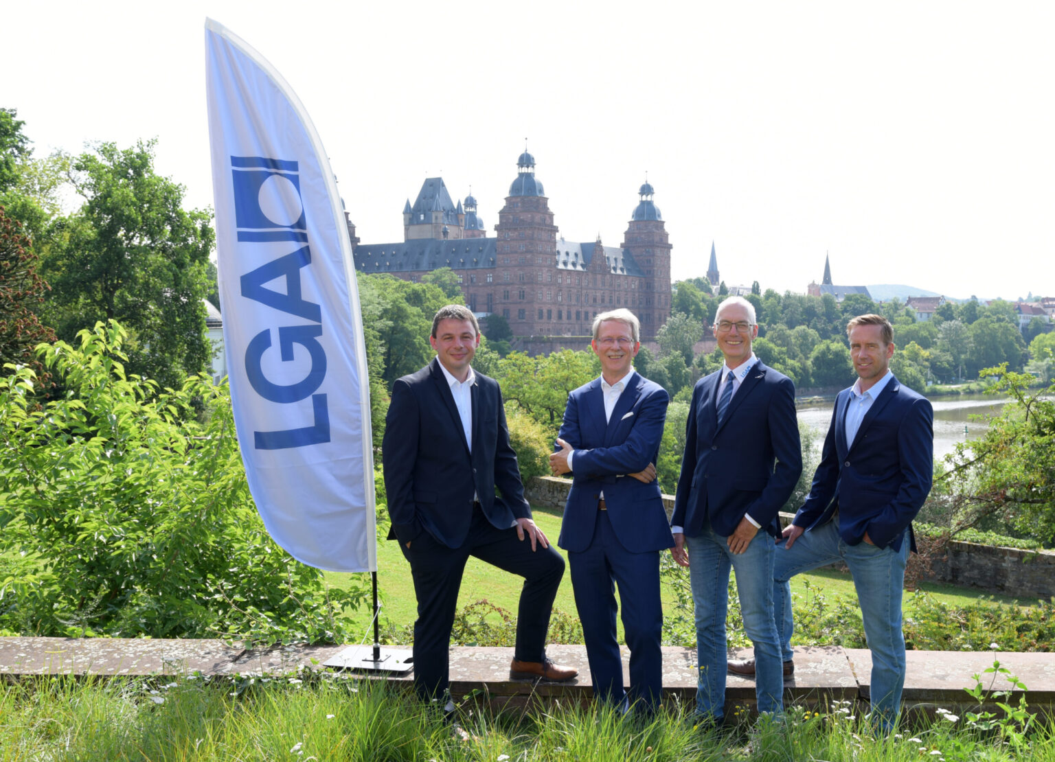 Vier Personen in formeller Kleidung stehen im Freien vor einem historischen Gebäude mit mehreren Türmen; links im Bild ist eine Fahne mit dem Schriftzug „LGA“ zu sehen, im Hintergrund befinden sich Bäume und Grünflächen.