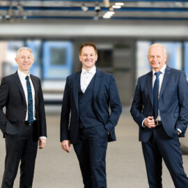 Drei Personen in formeller Businesskleidung stehen in einem großzügigen, modern gestalteten Innenraum.