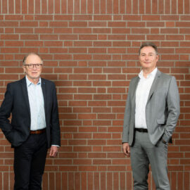 Zwei Personen in formeller Kleidung stehen vor einer Ziegelmauer; eine trägt einen dunklen Anzug mit hellblauem Hemd, die andere einen hellgrauen Anzug mit weißem Hemd.