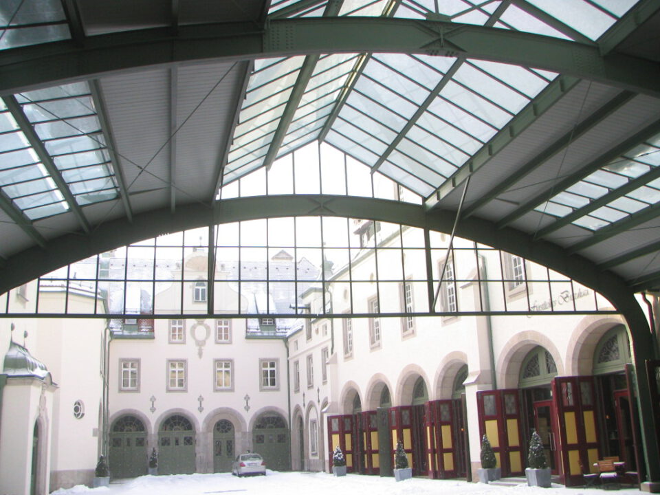 Geschlossener Innenhof mit Glasdach, umgeben von Gebäuden mit Rundbogentüren und -fenstern, schneebedecktem Boden und einem geparkten Auto in der Mitte des Hofes.
