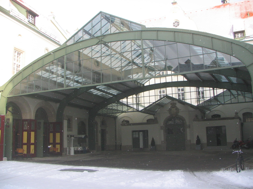 Großer, überdachter Innenhof mit einem gewölbten Glas- und Metalldach, umgeben von Gebäuden mit Rundbogentüren und -fenstern, während außerhalb der Überdachung Schnee liegt.