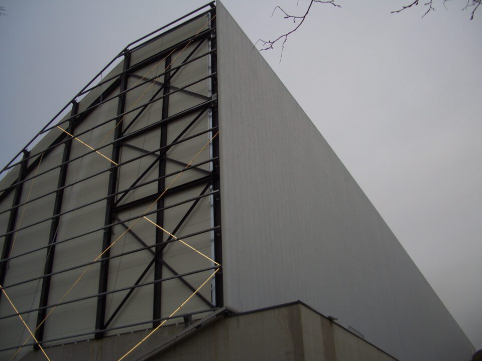 Großes, modernes Gebäude mit auffälliger Fassade aus schwarzen Metallträgern und weißen Paneelen, durchzogen von diagonalen Stützbalken; im Hintergrund ein bewölkter Himmel und Baumäste am oberen rechten Bildrand.