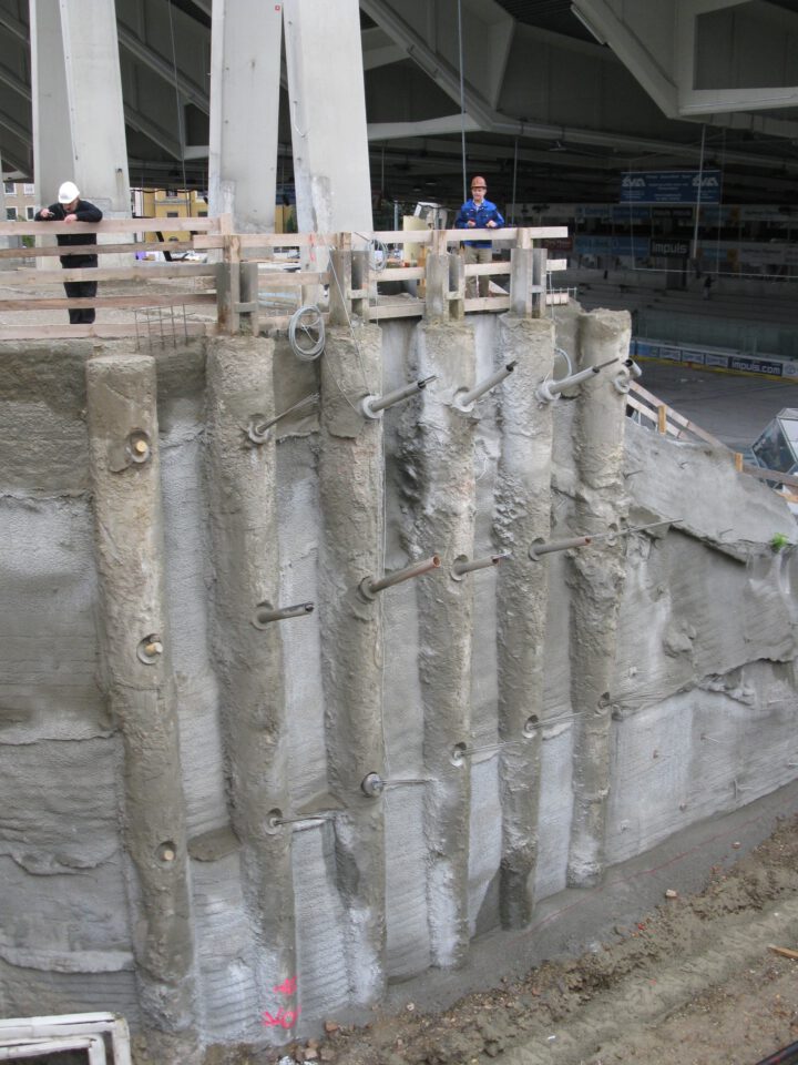 Baustelle mit einer Stützwand aus Betonpfählen, in die mehrere horizontale Metallstäbe eingelassen sind; links eine Person mit weißem Bauhelm, rechts eine weitere in blauer Arbeitskleidung, im Hintergrund Strukturelemente eines großen Hallenbaus.