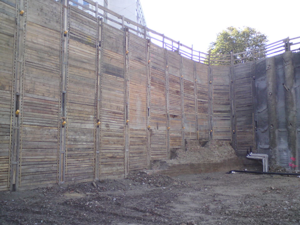 Baustelle mit großer Stützwand aus Holzbohlen und Metallverstrebungen, die sich bogenförmig um das Gelände zieht; am Fuß der Wand ist freigelegtes Erdreich sichtbar.