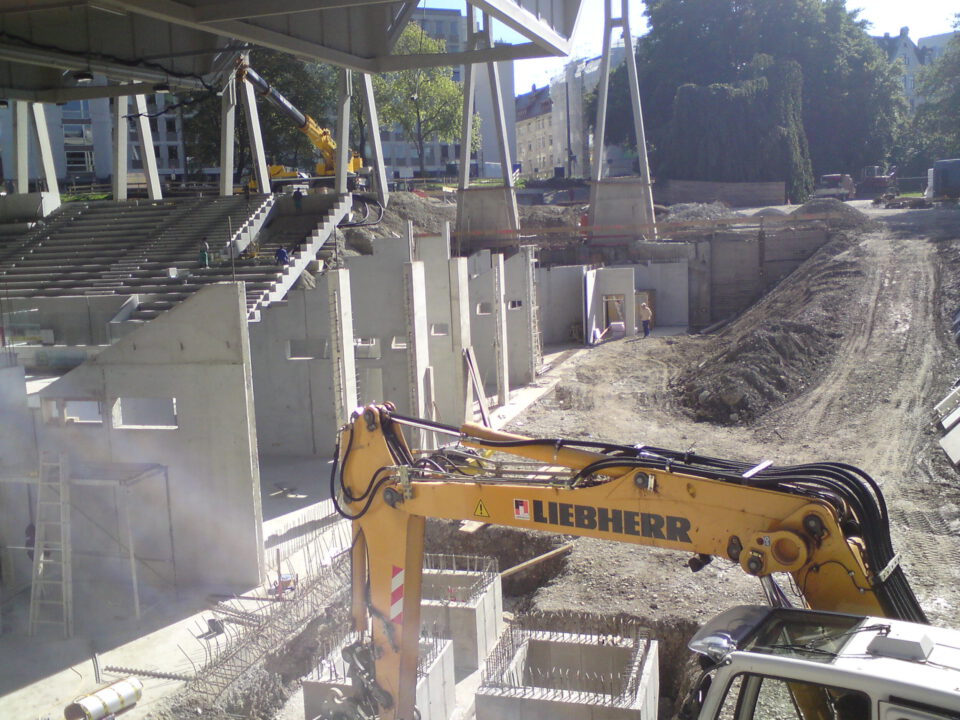 Baustelle mit großem gelbem LIEBHERR-Bagger, Betonbauteilen, Bewehrungsstahl und teils errichteten Wänden; eine Treppe führt zu einer erhöhten Plattform auf massiven Pfeilern, im Hintergrund Bäume und städtische Bebauung.