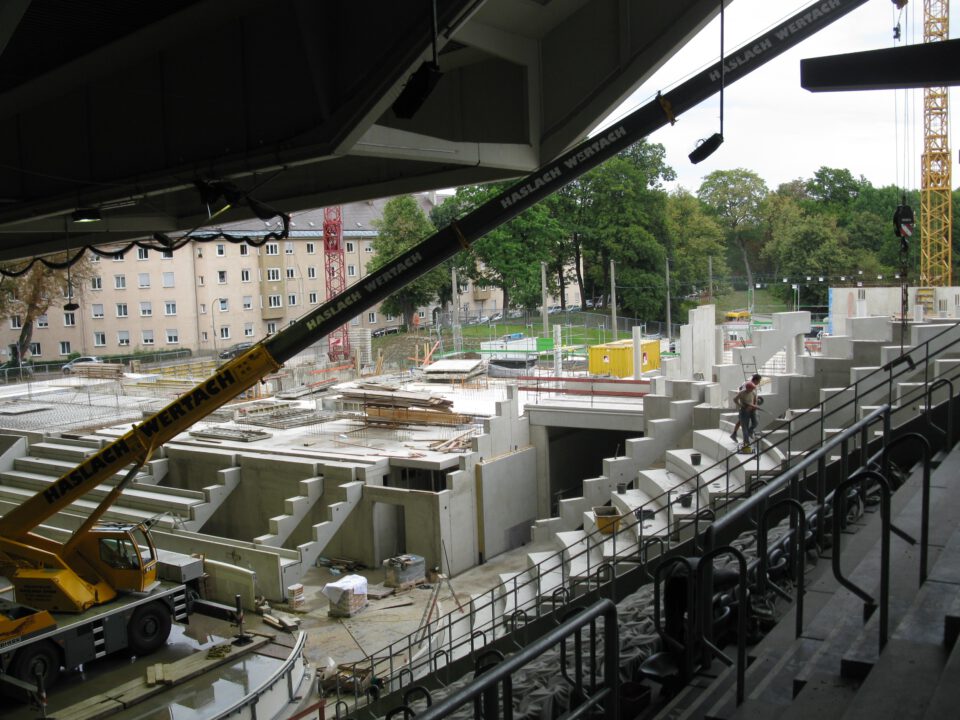 Baustelle eines großen öffentlichen Gebäudes mit Betonstufen und Gerüsten, im Vordergrund ein gelber Kran im Hintergrund Wohnhäuser und Bäume.