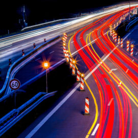 Das Bild zeigt eine nächtliche Autobahnszene mit roten und gelben Lichtspuren vorbeifahrender Fahrzeuge. Verkehrskegel, Absperrungen und ein Tempo-60-Schild wegen Bauarbeiten und Fahrbahnverengung.