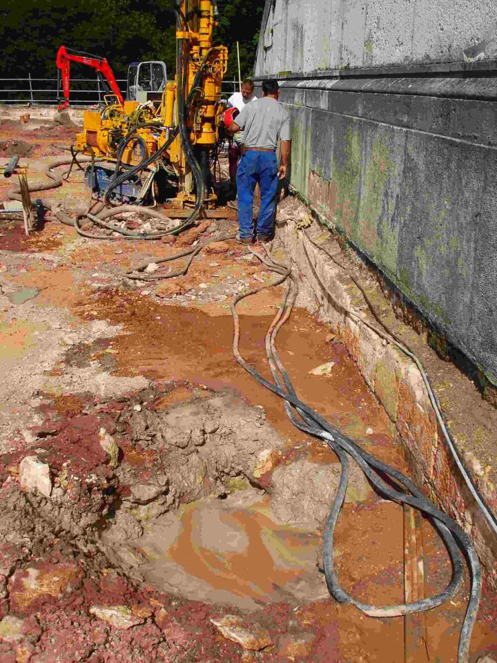 Auf einer Baustelle bedienen zwei Arbeiter eine große gelbe Bohrmaschine neben einer Betonwand, umgeben von Kabeln, Schläuchen, Erde, Steinen und Wasserpfützen
