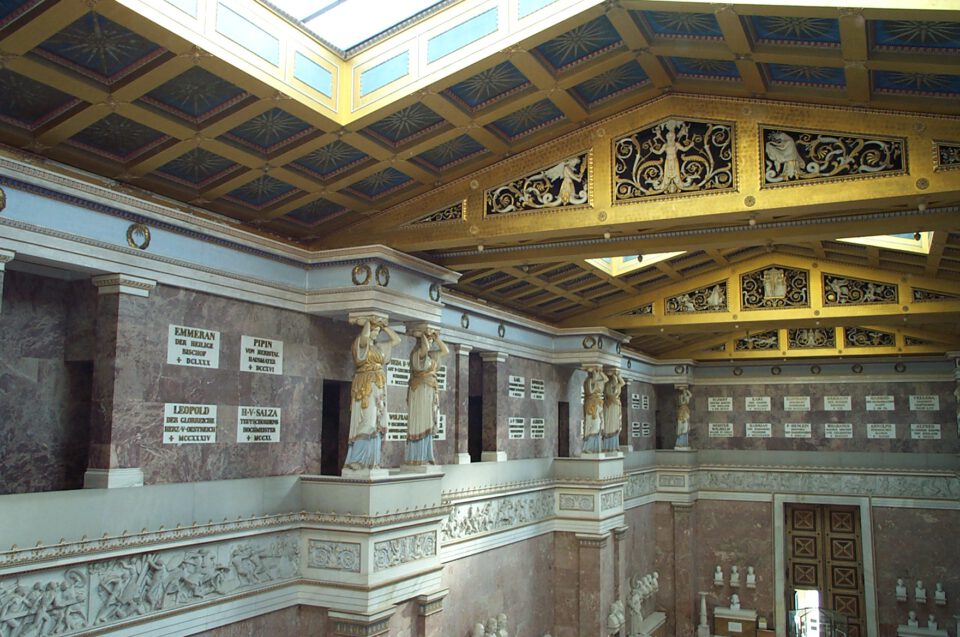 Das Bild zeigt das Innere einer prunkvollen Halle mit klassischer Architektur, reich verziertem goldenen Deckenschmuck, Statuen in antiker Kleidung und Wänden mit Namensplaketten und Inschriften.