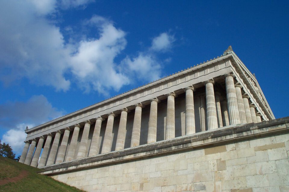 Das Bild zeigt ein großes klassizistisches Gebäude aus Stein mit hohen, kannelierten Säulen und einem dreieckigen Giebelfeld unter einem leicht bewölkten Himmel.