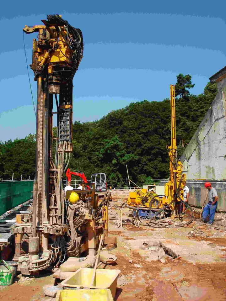 Das Bild zeigt eine Baustelle mit zwei großen Bohrgeräten, verstreutem Baumaterial und zwei Arbeitern, einer mit rotem Helm, vor einem Hintergrund aus Bäumen und einer teilweise sichtbaren Gebäudewand.