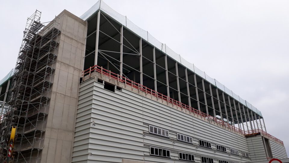 Großes Gebäude im Bau mit mehreren Etagen, links mit Gerüsten und oben mit offenem Tragwerk; der untere Bereich ist mit Metallverkleidung und mehreren Fenstern versehen, Sicherheitsnetze und Absperrungen kennzeichnen die laufenden Bauarbeiten.