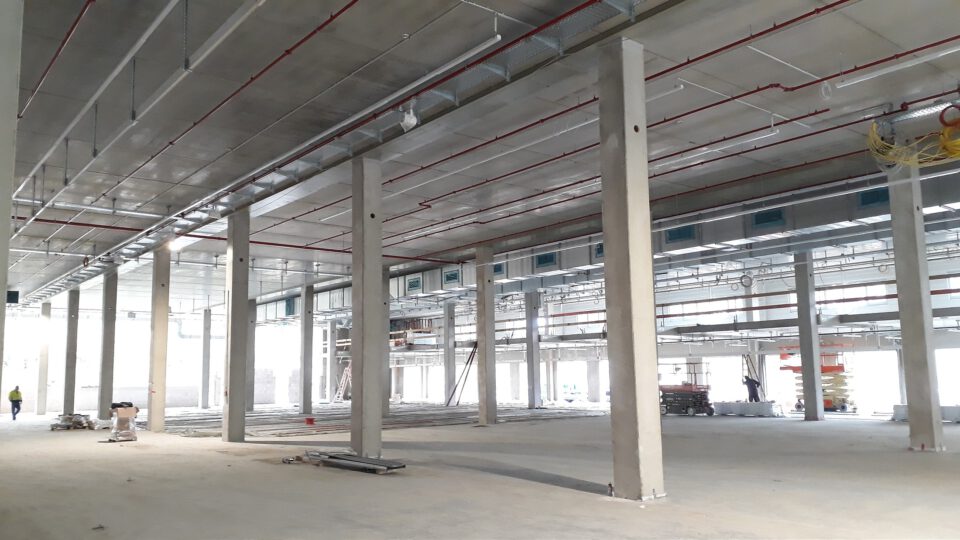 Innenraum eines großen, unfertigen Industrie- oder Gewerbebaus mit zahlreichen Betonsäulen, sichtbaren Rohrleitungen und Lüftungskanälen an der Decke sowie verstreuten Baumaterialien auf dem Boden.