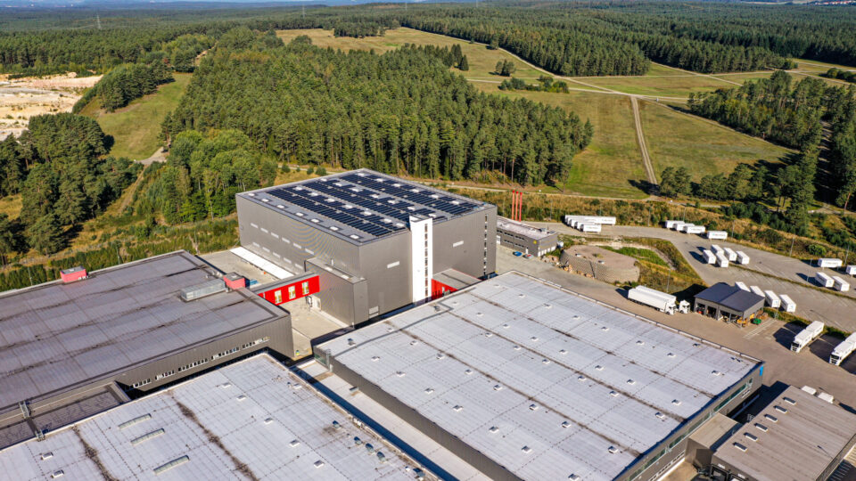 Luftaufnahme eines Industriekomplexes mit mehreren großen Flachdachgebäuden, teils mit Solarpanelen ausgestattet, umgeben von Wald und offenen Feldern; rund um die Gebäude sind zahlreiche Anhänger und Fahrzeuge geparkt, im Hintergrund dichter Wald und hügelige Landschaft.