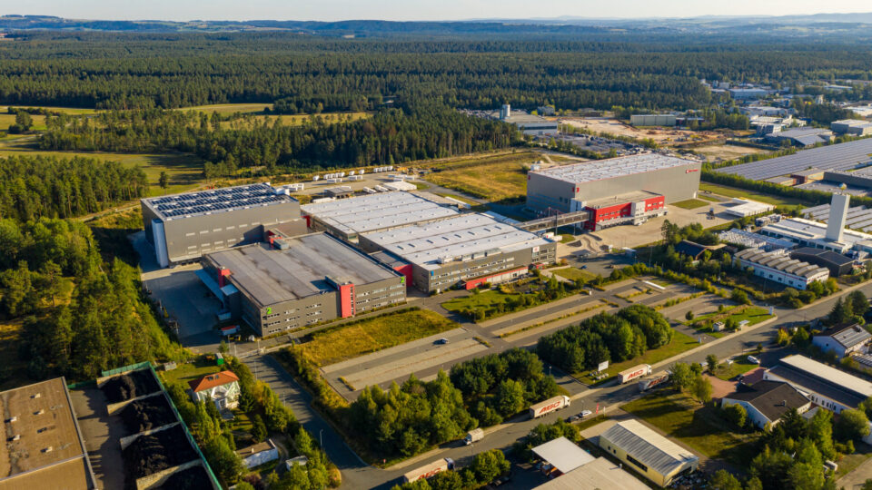 Luftaufnahme eines großen Industriekomplexes mit mehreren Gebäuden mit weißen und grauen Dächern sowie roten Akzenten, umgeben von Grünflächen, Wäldern und weiteren Industrie- oder Gewerbebauten; mehrere Lkw und Straßen verbinden die einzelnen Bereiche.