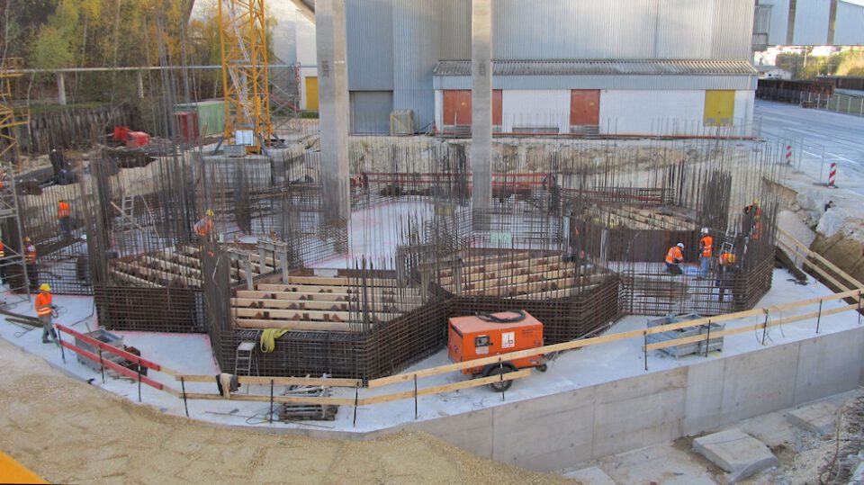 Baustelle mit mehreren Arbeitern in orangefarbener Schutzkleidung, Stahlbewehrungen und Schalungen im Rohbau, einem orangefarbenen Aggregat sowie Industriegebäuden mit Metallfassaden und bunten Toren im Hintergrund.