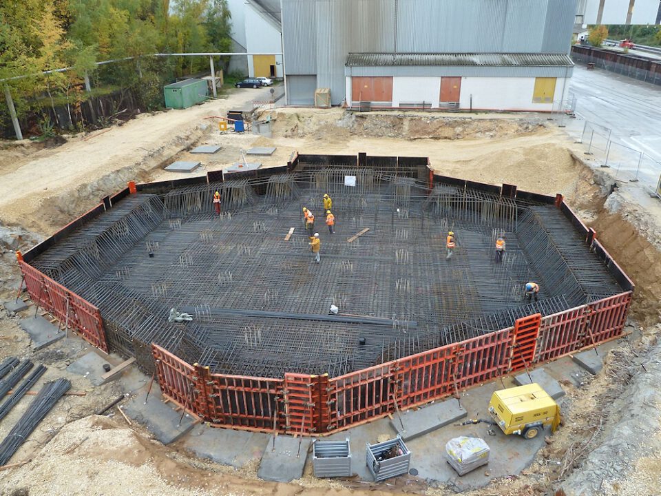 Baustelle mit mehreren Arbeitern in Schutzkleidung, einer achteckigen Fundamentstruktur mit Stahlbewehrung und roten Schalungselementen, umgeben von Baumaterialien und einem Industriegebäude im Hintergrund.