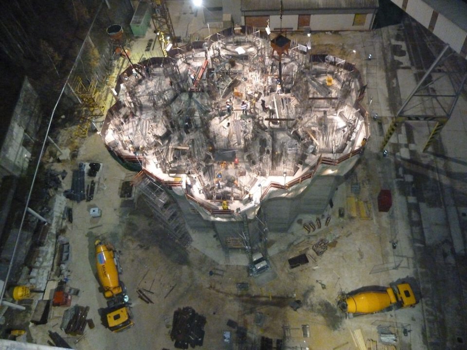 Nächtliche Baustelle mit einem großen, beleuchteten Wabenförmigen im Zentrum, umgeben von Baugeräten, Gerüsten, Baumaterialien und zwei gelben Betonmischern im Vordergrund.