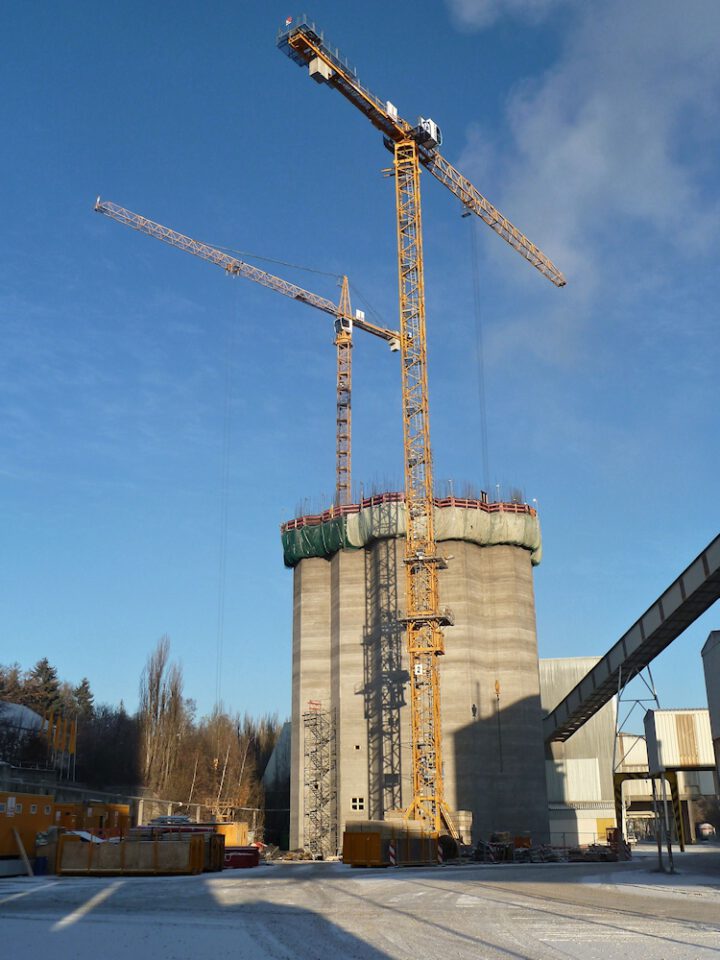 Baustelle mit zwei großen Kränen, die an einem hohen zylindrischen Betonbau mit Gerüst und grüner Netzeinfassung arbeiten, umgeben von Containern, kleineren Gebäuden und Bäumen unter leicht bewölktem Himmel.