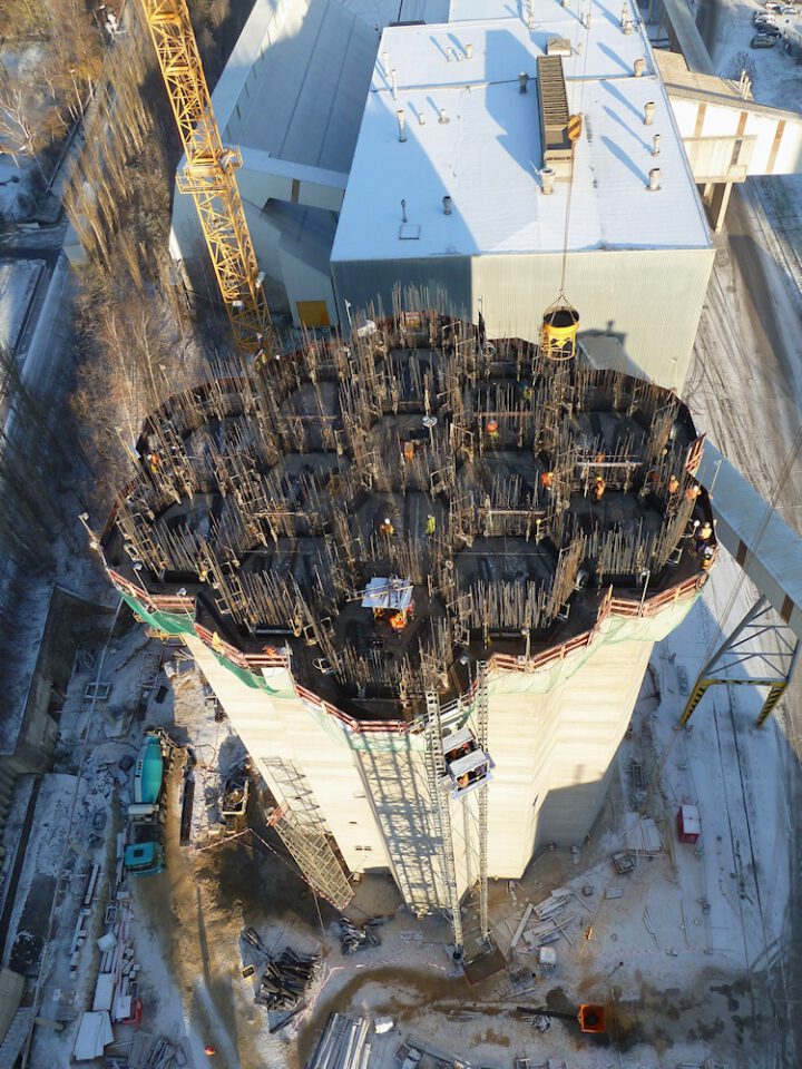 Luftaufnahme einer Baustelle mit einem großen zylindrischen Bauwerk, mehreren wabenförmigen Segmenten an der Oberseite, Gerüsten, Bewehrung, einem Kran sowie sichtbaren Bauarbeitern und umliegenden Materialien und Gebäuden