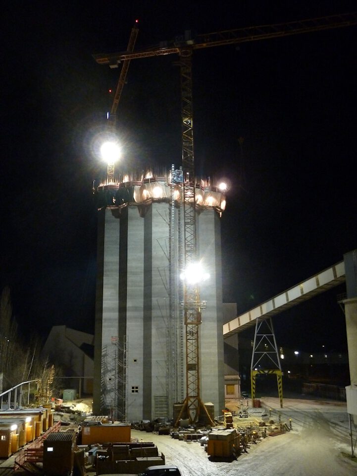 Baustelle bei Nacht mit einem hohen zylindrischen Bauwerk im Bau, zwei großen Kränen und heller Beleuchtung, umgeben von Baumaterialien und Baumaschinen.