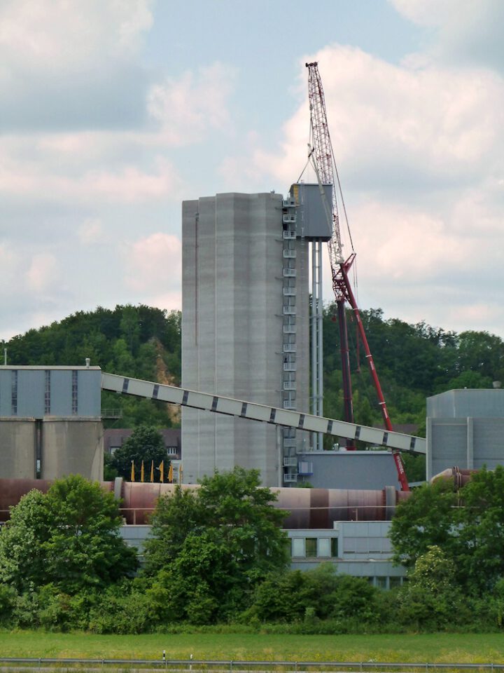 Industrieanlage mit einem hohen rechteckigen Betonbau im Zentrum, einem roten Kran und einem Förderband, umgeben von Bäumen, Wiesen und Hügeln unter teils bewölktem Himmel.