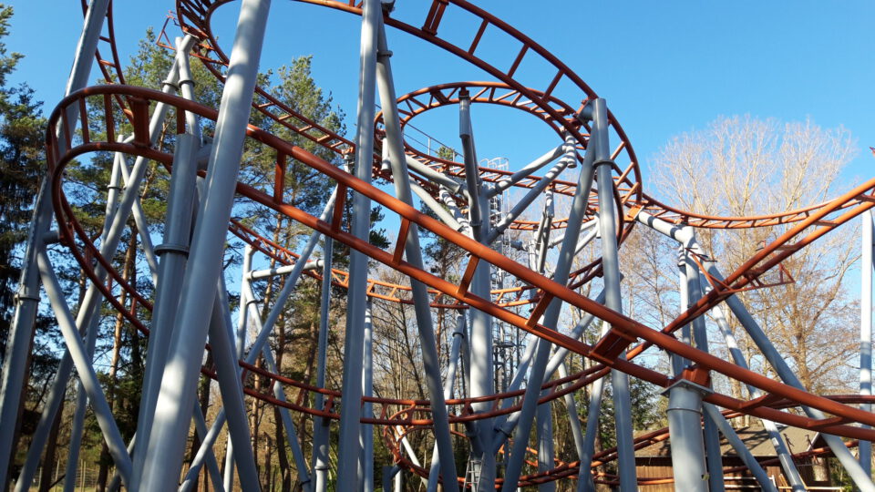 Komplexe Achterbahn mit orangefarbenen Schienen und grauen Metallstützen, mehreren Loopings und Kurven, vor einem Hintergrund aus Bäumen und blauem Himmel.