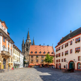 Stadtplatz in Ansbach mit farbenfrohen historischen Gebäuden, einem zentralen Bau mit rotem Ziegeldach und Gaubenfenstern, umgeben von Häusern mit verzierten Fassaden, Blumen geschmückten Fenstern und Außensitzplätzen unter Sonnenschirmen bei sonnigem Wetter.