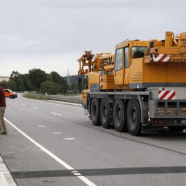 Das Bild zeigt eine Szene auf einer Landstraße mit einem großen gelben Kranfahrzeug, das am rechten Fahrbahnrand steht. Vier Personen sind zu sehen, darunter jemand mit orangefarbener Warnweste und weißem Helm sowie weitere Personen in Arbeits- oder Freizeitkleidung. Im Hintergrund befinden sich Bäume, Felder und ein bewölkter Himmel.
