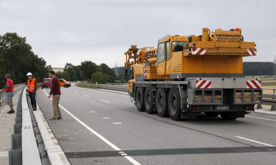 Das Bild zeigt eine Szene auf einer Landstraße mit einem großen gelben Kranfahrzeug, das am rechten Fahrbahnrand steht. Vier Personen sind zu sehen, darunter jemand mit orangefarbener Warnweste und weißem Helm sowie weitere Personen in Arbeits- oder Freizeitkleidung. Im Hintergrund befinden sich Bäume, Felder und ein bewölkter Himmel.