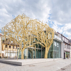 Das Bild zeigt eine moderne architektonische Struktur mit einem auffälligen Design, das an einen stilisierten Baum aus gelben Metallträgern erinnert. Das Gebäude verfügt über große Glasflächen und befindet sich inmitten traditioneller Gebäude in einem städtischen Umfeld. Es wirkt wie ein öffentlich zugänglicher oder kommerziell genutzter Raum.