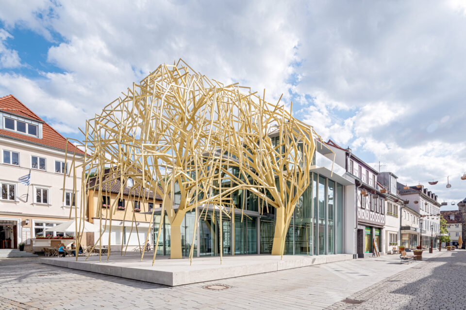 Das Bild zeigt eine moderne architektonische Struktur mit einem auffälligen Design, das an einen stilisierten Baum aus gelben Metallträgern erinnert. Das Gebäude verfügt über große Glasflächen und befindet sich inmitten traditioneller Gebäude in einem städtischen Umfeld. Es wirkt wie ein öffentlich zugänglicher oder kommerziell genutzter Raum.