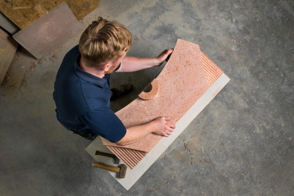 Eine Person arbeitet mit einem großen Stück Naturstein, das eine gebogene Form und eine strukturierte Oberfläche hat. Auf dem Boden liegen Werkzeuge wie Hammer und Meißel. Die Szene deutet auf handwerkliche Bearbeitung von Naturstein für Bauzwecke hin.