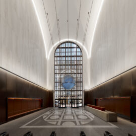 Das Bild zeigt eine moderne, großzügige Eingangshalle mit hohen Decken und einem großen Rundbogenfenster am Ende. Die Wände bestehen aus hellem Marmor, in der Mitte des Bodens befindet sich ein geometrisches Muster. Vor dem Fenster schwebt eine blaue Kugelskulptur, und an der Decke verlaufen LED-Lichtstreifen. Am Eingang sind mehrere Zugangsschranken zu sehen.